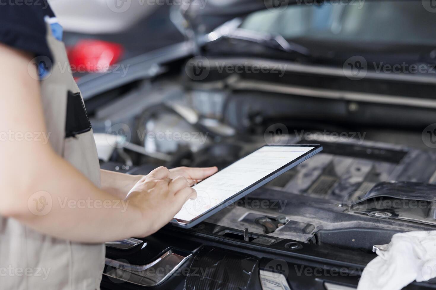 Certified mechanic in garage using tablet to follow checklist while doing maintenance on car. Close up shot of specialist in auto repair shop doing checkup on vehicle engine photo