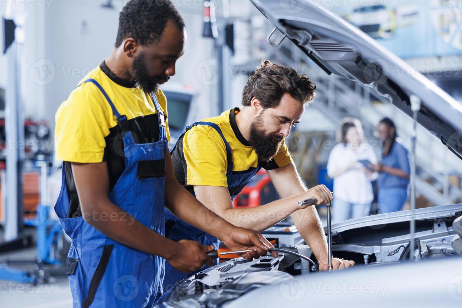 Coworkers using oil extractor tool to change old depleted brake fluid with new one in auto repair shop. BIPOC mechanic and colleague using professional equipment to do car maintenance photo