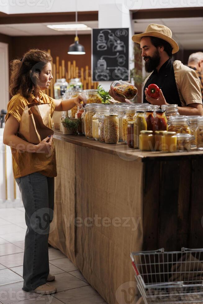Client shopping for organic pantry staples in zero waste store using biodegradable paper bag, being assisted with information by trader. Vendor showing woman products in reusable jars photo