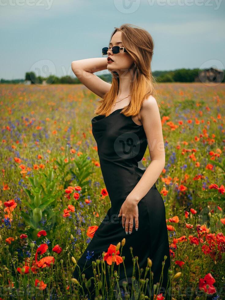 Beautiful young girl in a black evening dress and sunglasses posing against a poppy field on a cloudy summer day. Portrait of a female model outdoors. Rainy weather. Gray clouds. photo