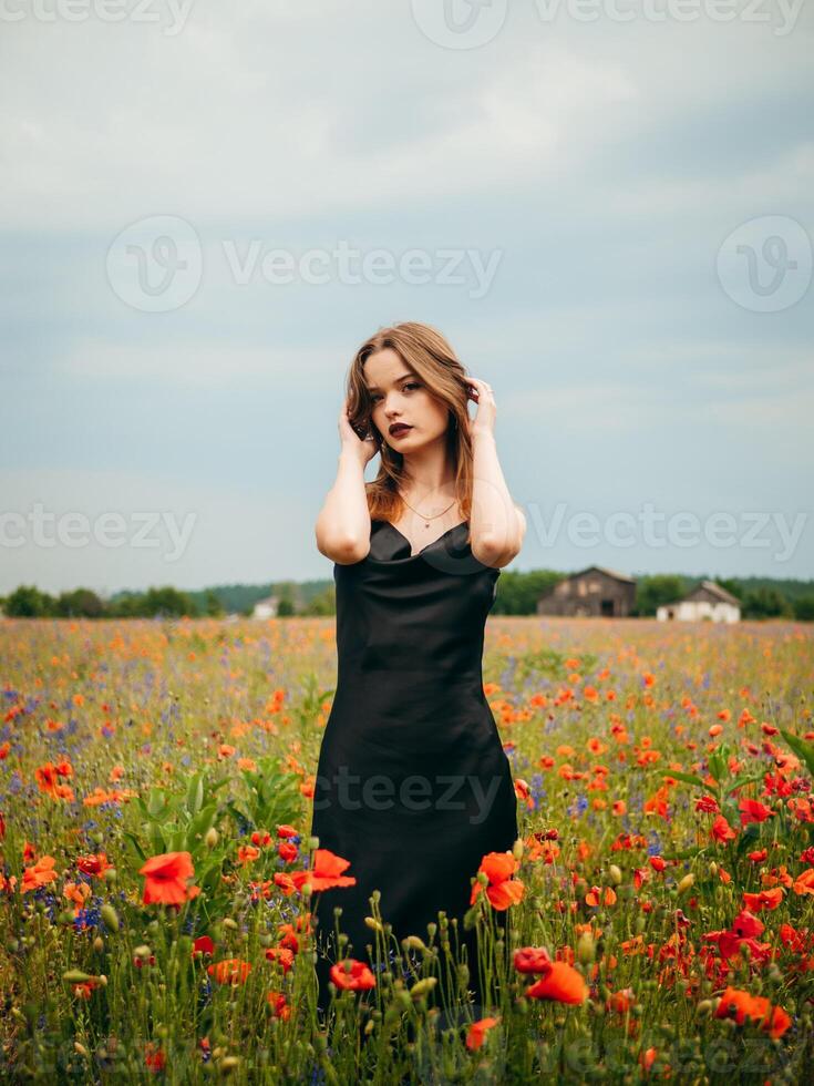 Beautiful young girl in a black evening dress posing against a poppy field on a cloudy summer day. Portrait of a female model outdoors. Rainy weather. Gray clouds. photo