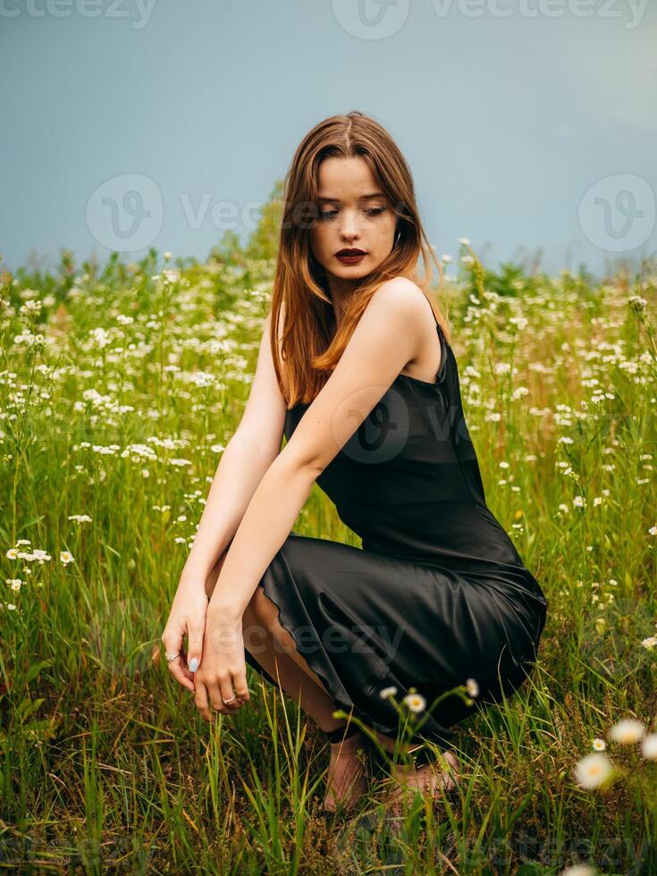 Beautiful young girl in a black evening dress poses crouching in front of a daisy field on a cloudy summer day. Portrait of a female model outdoors. Rainy weather. Gray clouds. photo