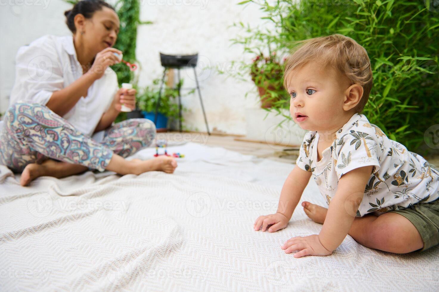 Curious cute baby boy in khaki color shorts and nature patterned white shirt, sitting on a blanket on the floor in the home garden. His mom is blowing soap bubbles on the blurred background. photo