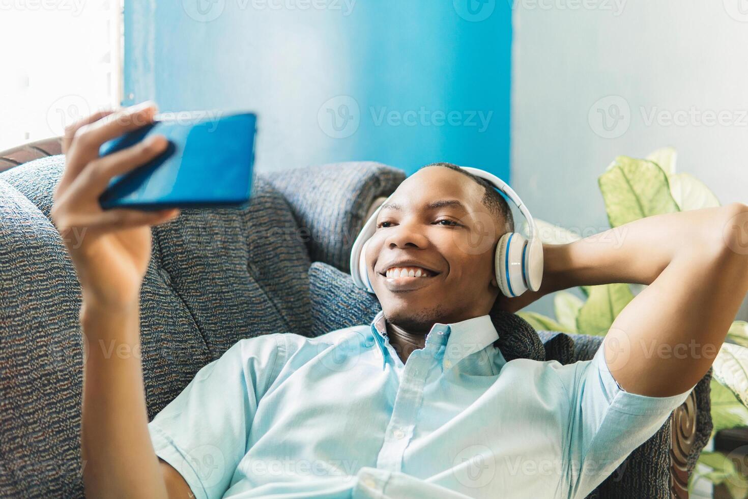 A happy african man listens and watches the content of the phone sitting on a sofa in the day at home. photo