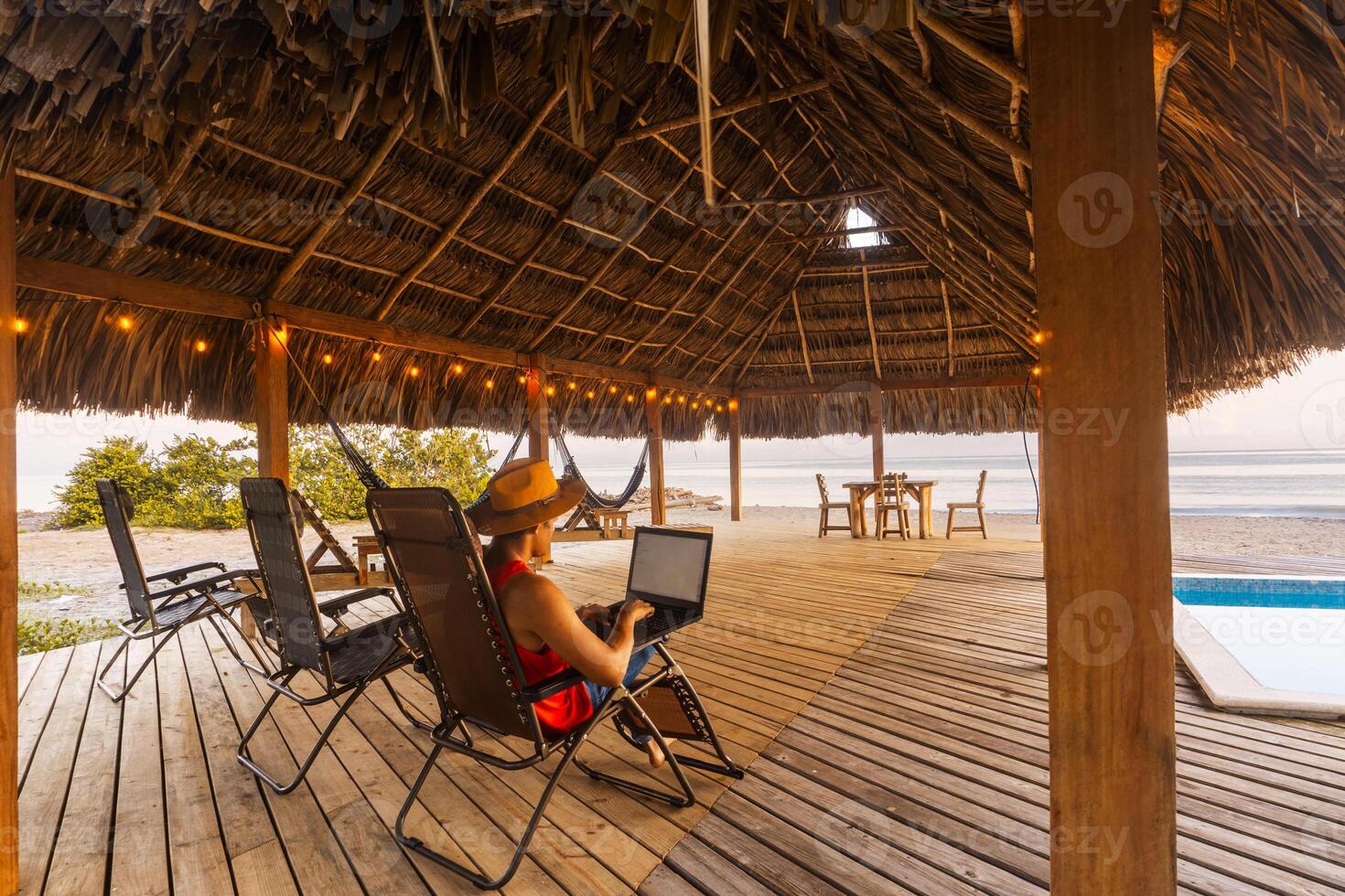 Man with a hat sitting on a chair and working on a laptop in front of the beach. photo