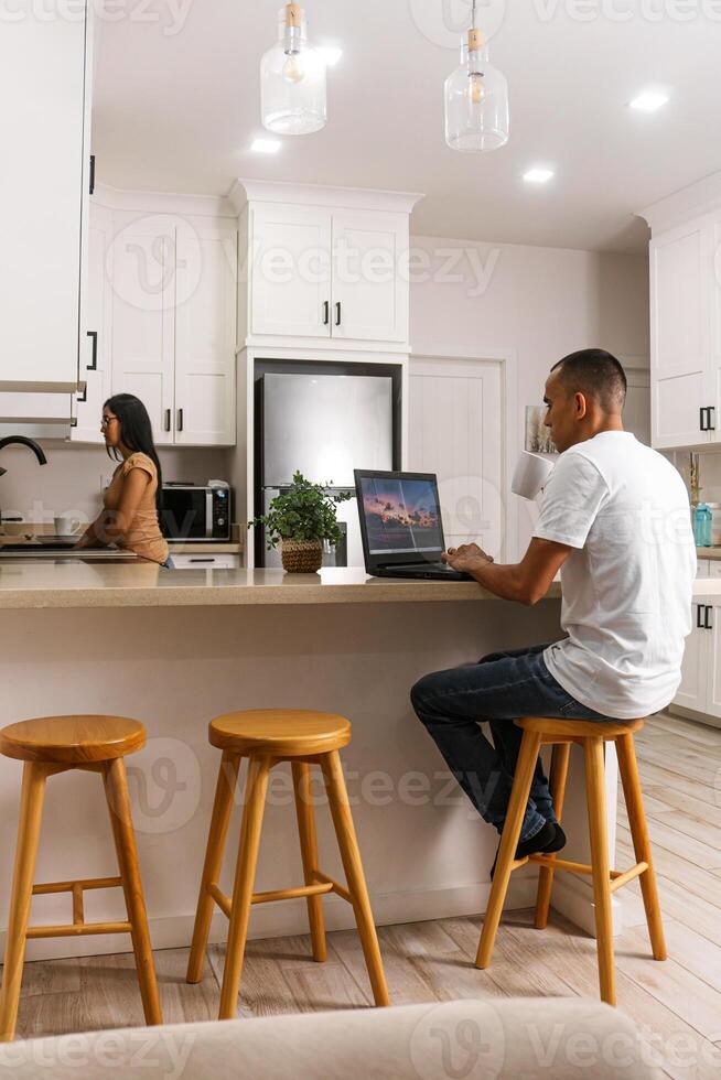 Young man working on his computer having a cup of a drink, in the background his partner doing the cleaning at the sink. photo