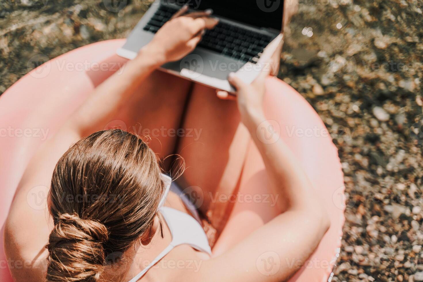 A woman is sitting in a pink inflatable pool with a laptop on her lap. She is typing on the laptop, possibly working or browsing the internet. Concept of relaxation and leisure. photo