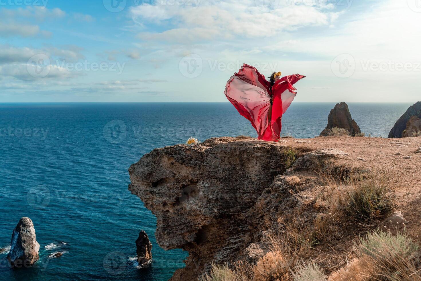 A woman in a red dress stands on a rocky cliff overlooking the ocean. The scene is serene and peaceful, with the woman's flowing dress adding a sense of grace and beauty to the landscape. photo