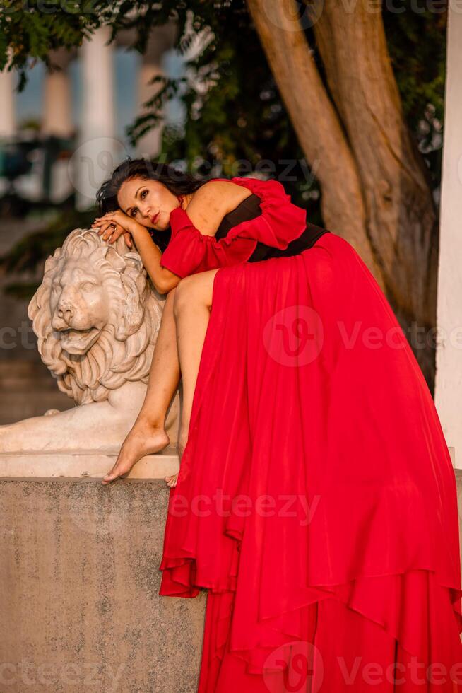 A woman in a red dress is sitting on a stone lion statue. The statue is white and has a lion's head. The woman is wearing a black belt and is looking at the camera. The scene has a calm. photo