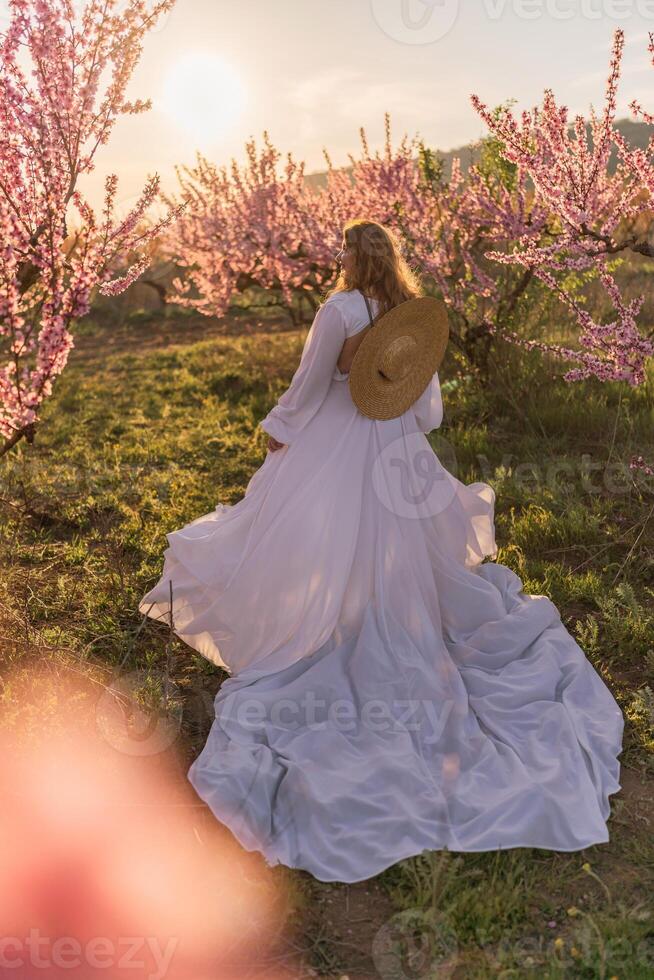 Woman blooming peach orchard. Against the backdrop of a picturesque peach orchard, a woman in a long white dress and hat enjoys a peaceful walk in the park, surrounded by the beauty of nature. photo