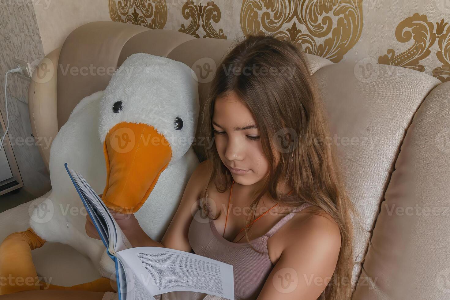 A girl is reading a book while sitting on a bed with a stuffed duck. The room has a gold and white color scheme, and there are two lamps in the room. photo