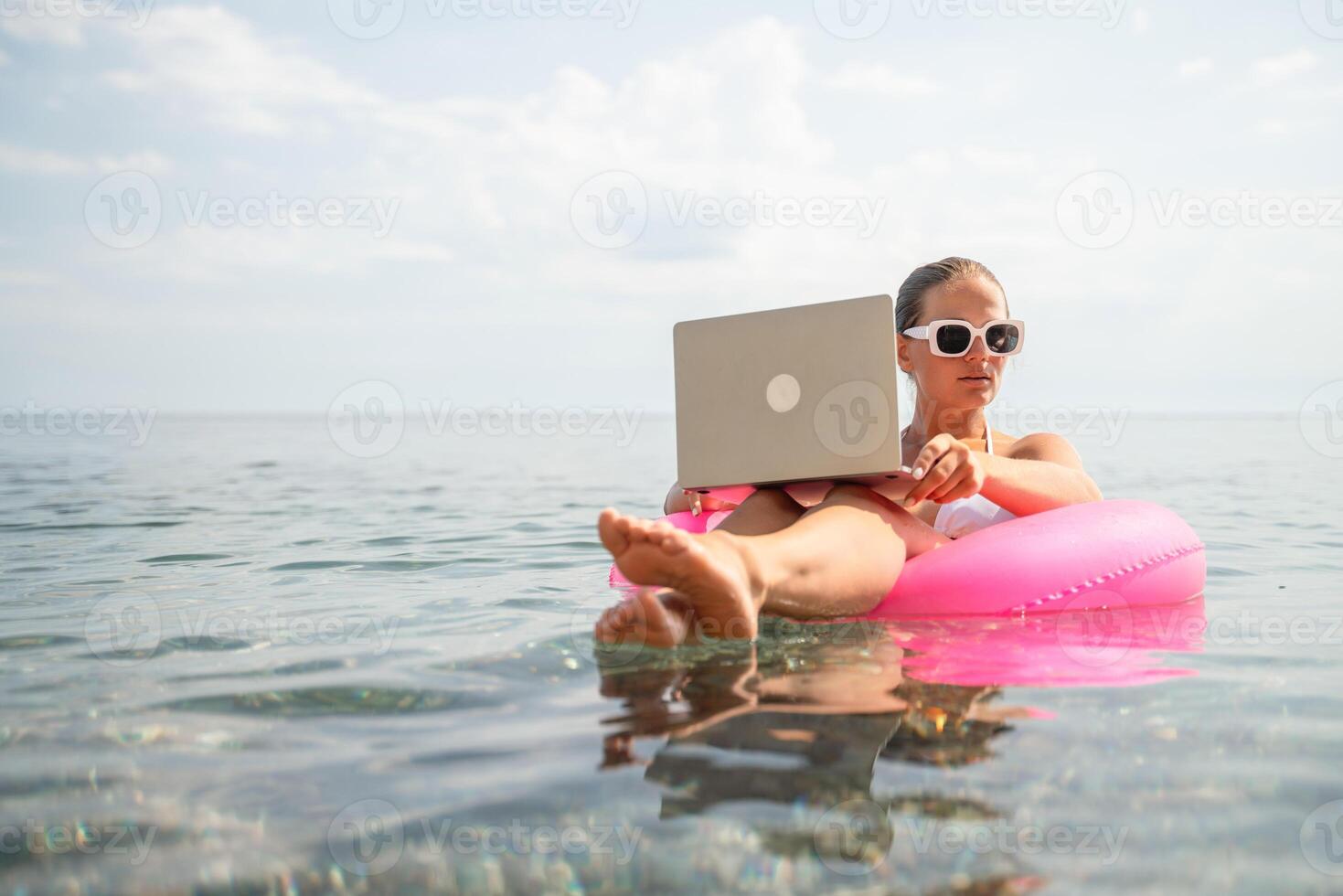 A woman is sitting on a pink inflatable raft in the ocean, using a laptop. Concept of relaxation and leisure, as the woman is enjoying her time in the water while working on her laptop. photo