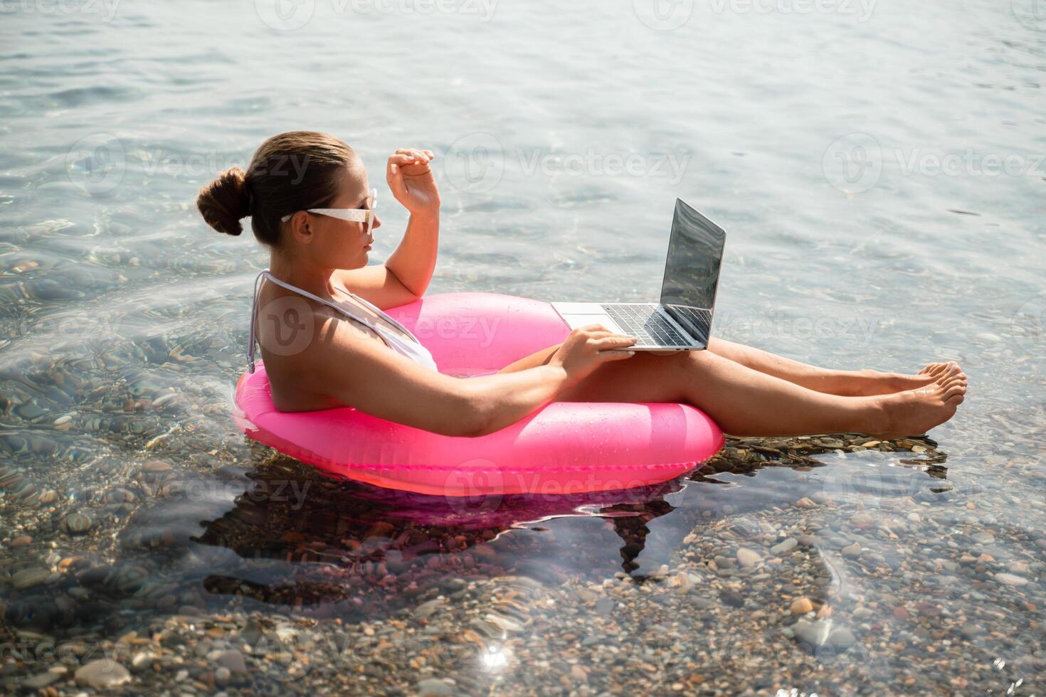 A woman is sitting on a pink inflatable raft in the ocean, using a laptop. Concept of relaxation and leisure, as the woman enjoys her time by the water while working on her laptop. photo