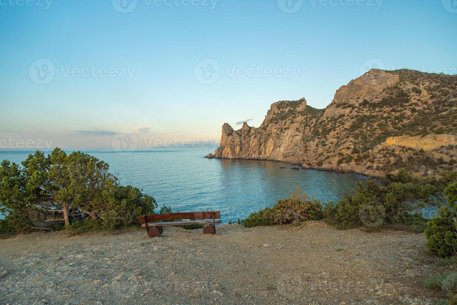 A bench is sitting on a rocky beach next to the ocean. The sky is blue and the sun is setting. photo