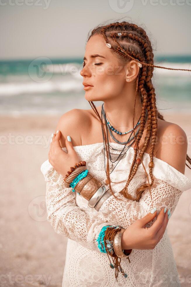 Model in boho style in a white long dress and silver jewelry on the beach. Her hair is braided, and there are many bracelets on her arms. photo