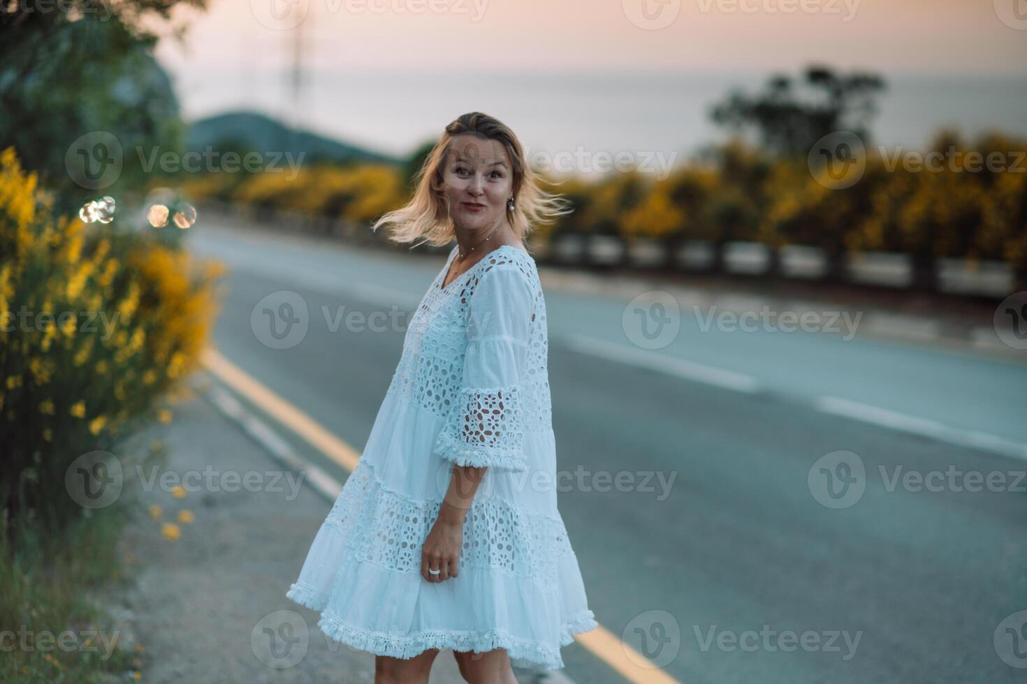 A woman in a white dress stands on a road next to a yellow bush photo