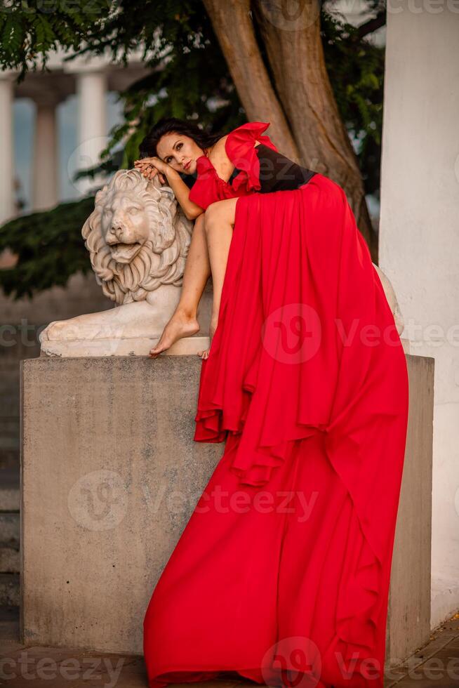 A woman in a red dress is sitting on a stone lion statue. The statue is white and has a lion's head. The woman is wearing a black belt and is looking at the camera. The scene has a calm. photo