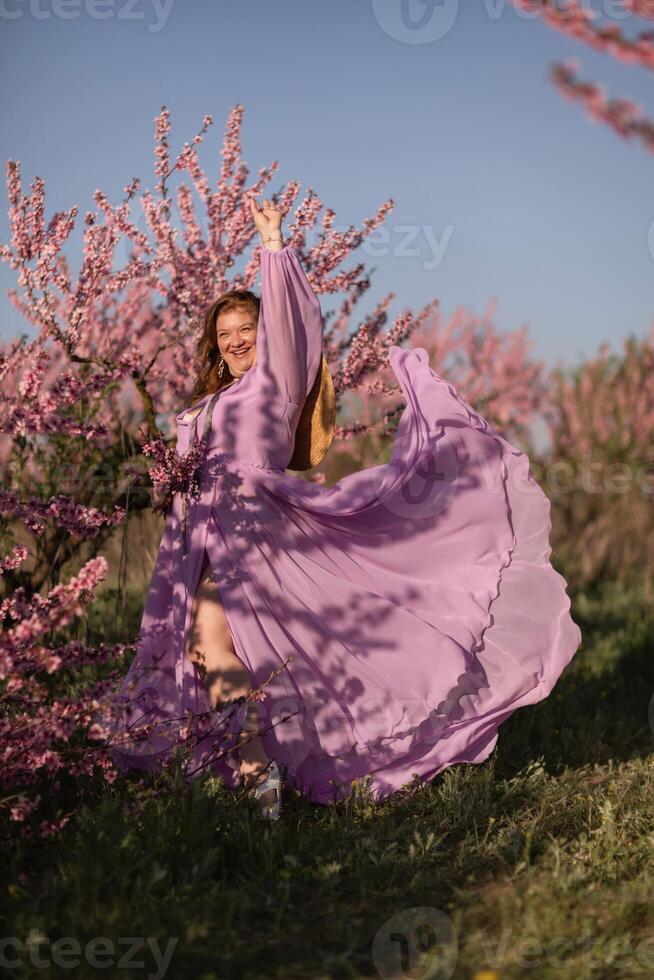 Woman blooming peach orchard. Against the backdrop of a picturesque peach orchard, a woman in a long pink dress and hat enjoys a peaceful walk in the park, surrounded by the beauty of nature. photo