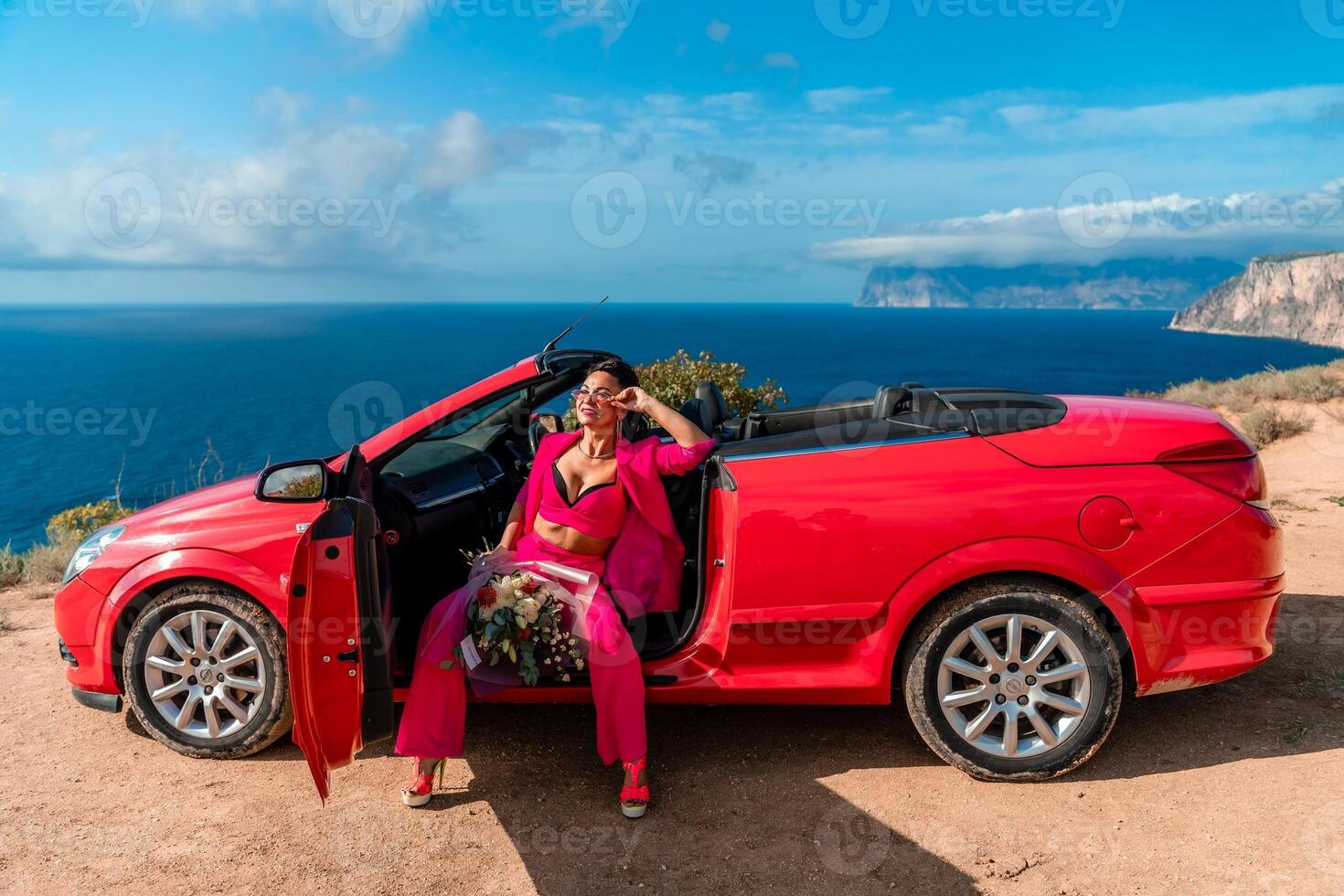 A woman is sitting in the back of a red convertible car. The car is parked on a dirt road near the ocean. The woman is wearing a pink suit and red shoes. Scene is relaxed and carefree. photo