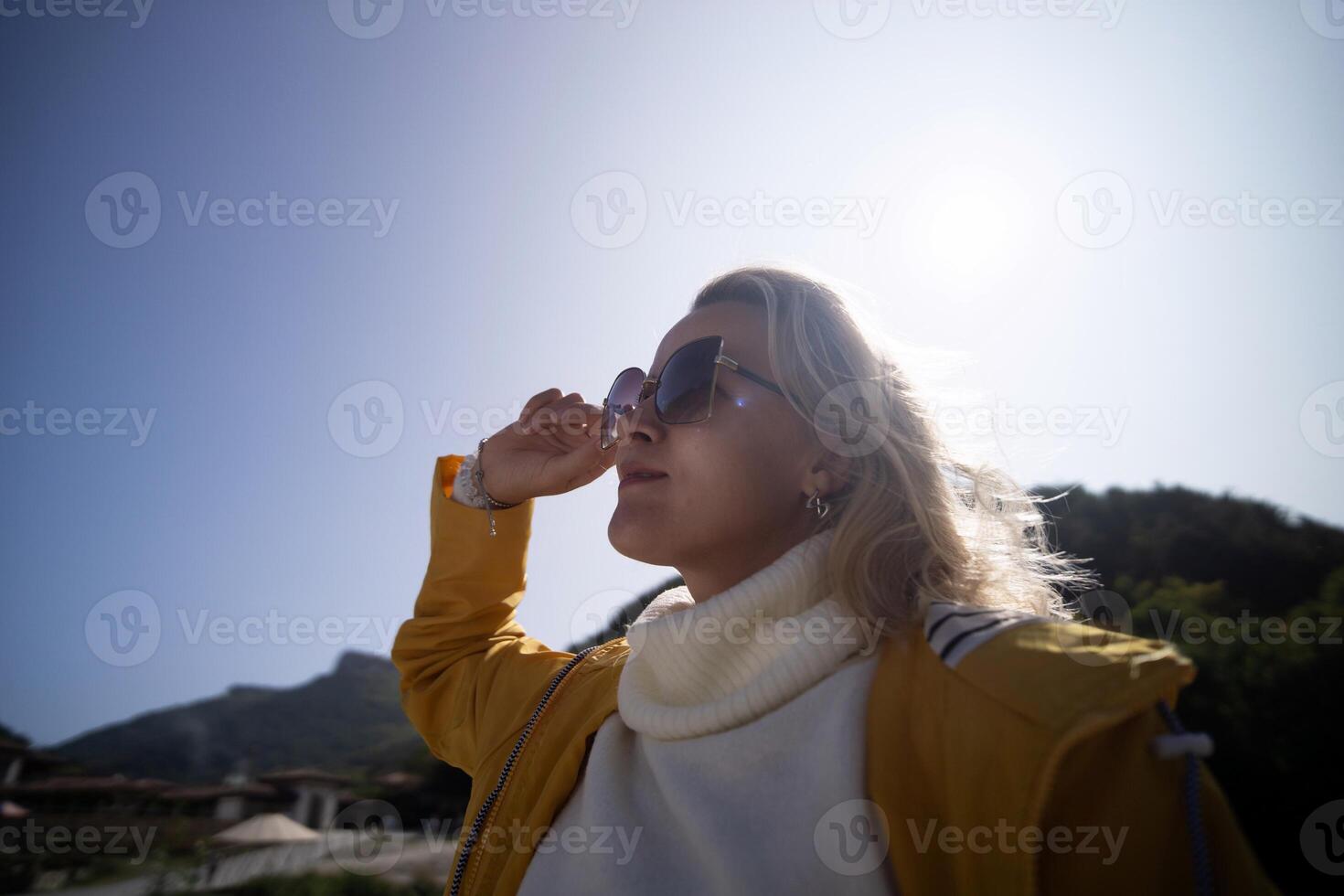 A woman wearing a yellow jacket and sunglasses is standing in the sun photo