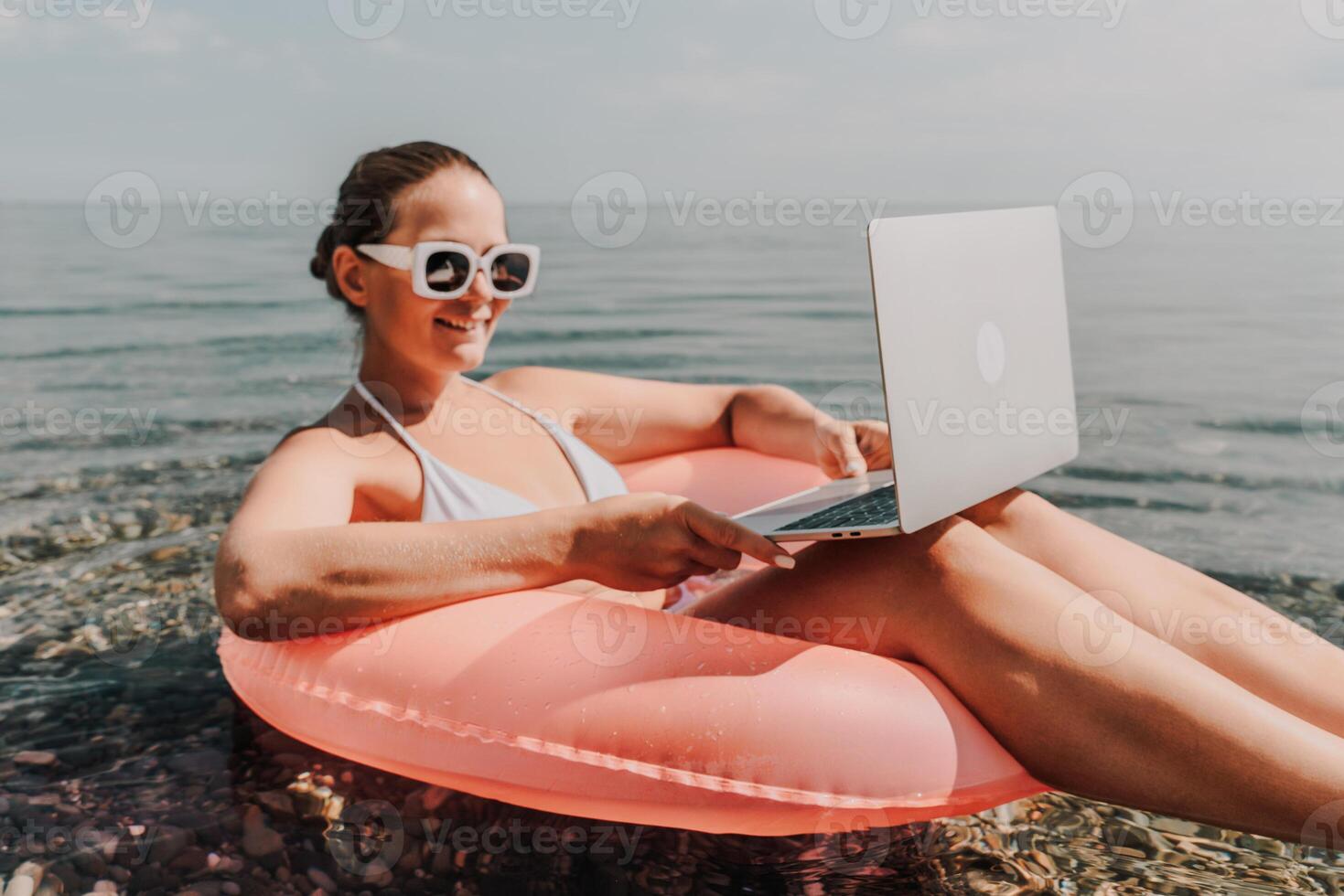 A woman is sitting on a pink inflatable raft with a laptop on her lap. She is wearing sunglasses and a white bikini top. Concept of relaxation and leisure. photo