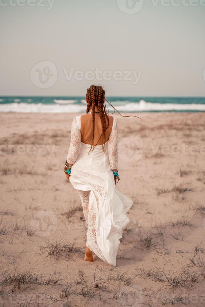 Model in boho style in a white long dress and silver jewelry on the beach. Her hair is braided, and there are many bracelets on her arms. photo