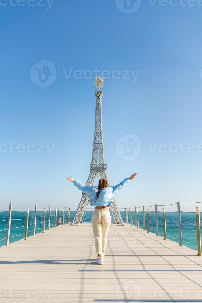 Large model of the Eiffel Tower on the beach. A woman walks along the pier towards the tower, wearing a blue jacket and white jeans. photo