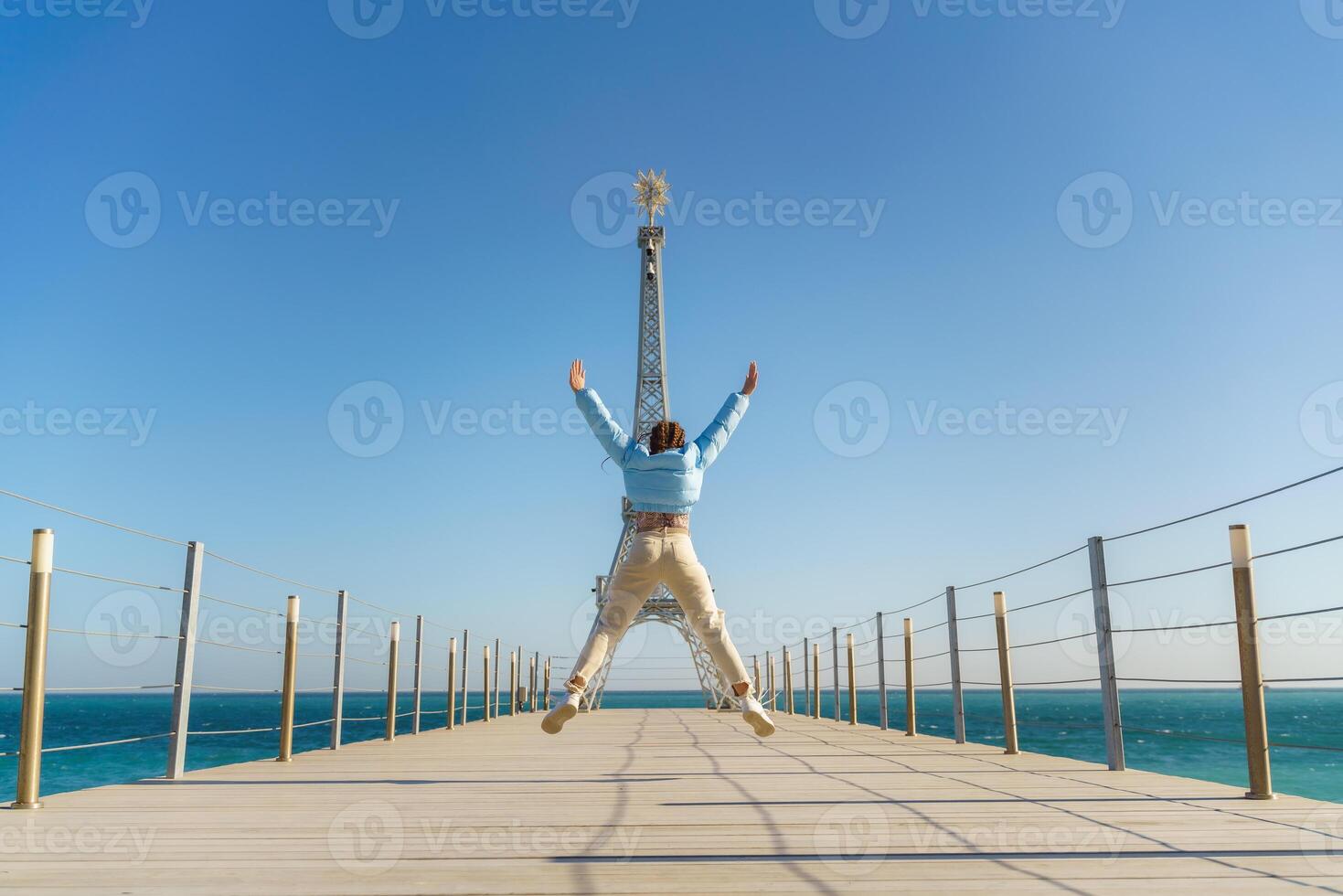 Large model of the Eiffel Tower on the beach. A woman walks along the pier towards the tower, wearing a blue jacket and white jeans. photo