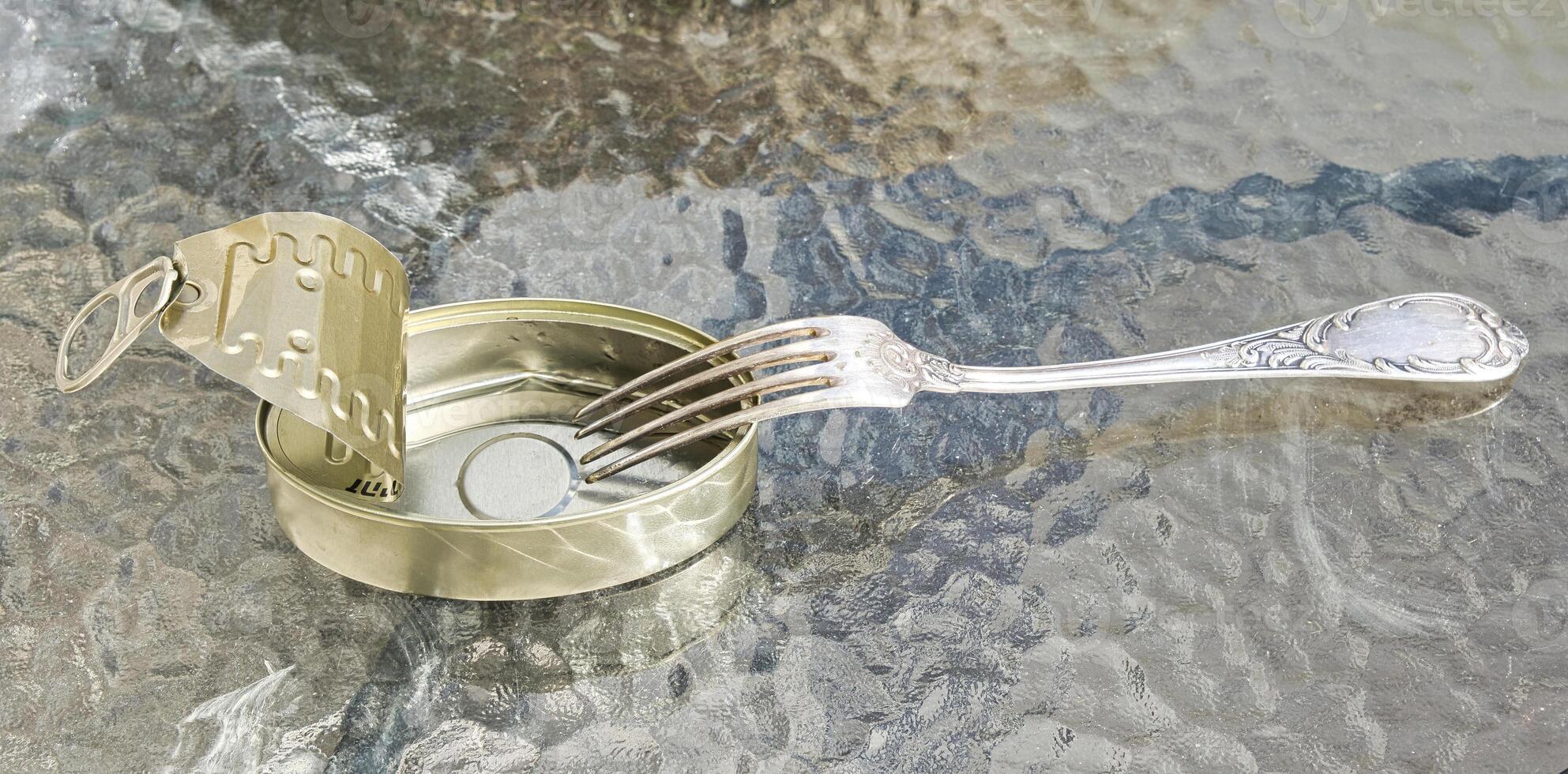 Open empty tin can and fork on the glass table surface. photo