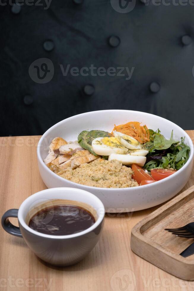 A bowl of couscous with meat, eggs and avocado, vegetables and a cup of coffee on the restaurant table photo