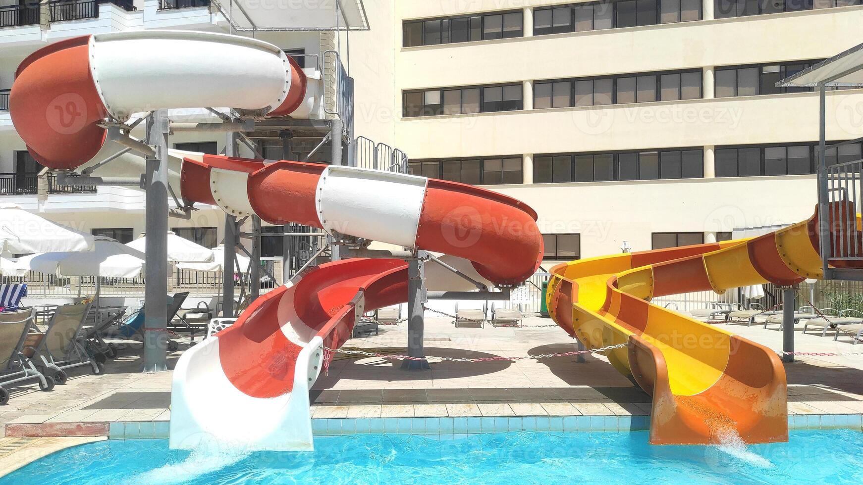 Colorful water slides at a resort water park, with red, orange, and yellow tubes leading into a refreshing pool. Sun loungers and umbrellas provide a relaxing atmosphere for a fun-filled summer day photo