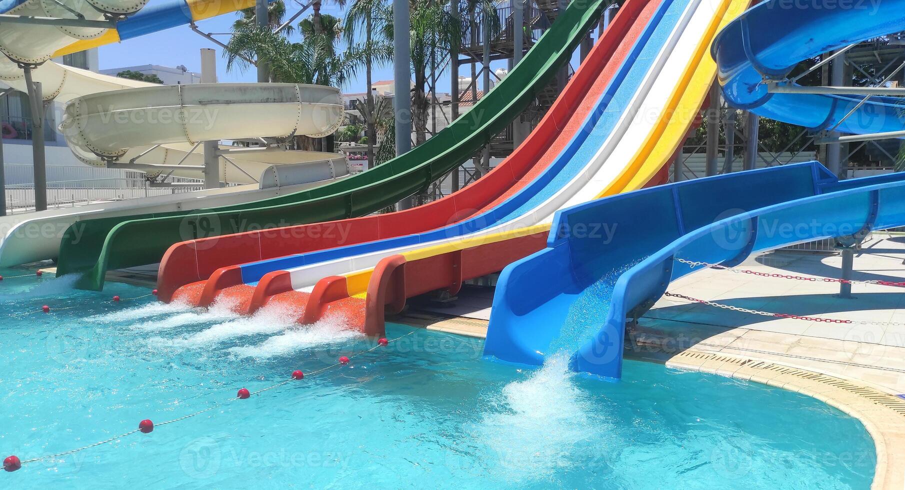 Sunny day at a lively water park with vibrant water slides and a splash pool. Surrounded by palm trees and under a clear blue sky, tropical vacation feel, ideal for family fun and excitement. photo
