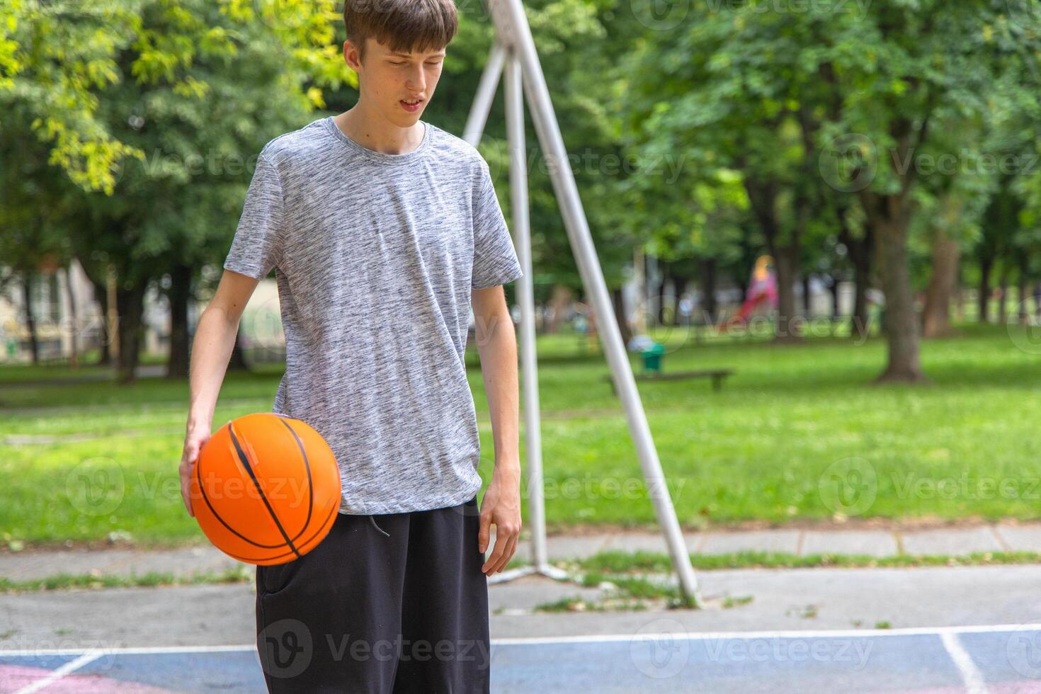 A teenage boy stands on an outdoor basketball court, holding a basketball in his left hand. He is wearing a grey t-shirt and black shorts. The court is surrounded by green grass and trees. photo