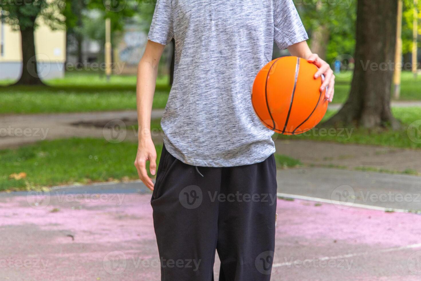 A young boy wearing a grey t-shirt and black pants stands on a basketball court, holding a basketball in his right hand. The court is surrounded by green grass, trees, and houses. The day is sunny. photo