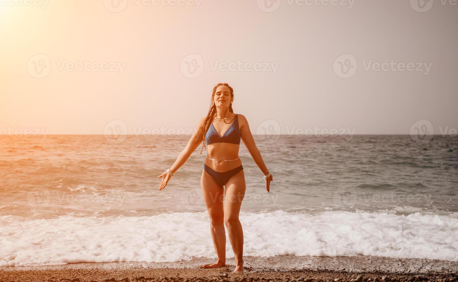 A tourist woman in blue bikini on beach enjoys the turquoise sea during her summer holiday. Rear view. Beach vacation. woman with braids dreadlocks standing with her arms raised enjoying beach ocean. photo