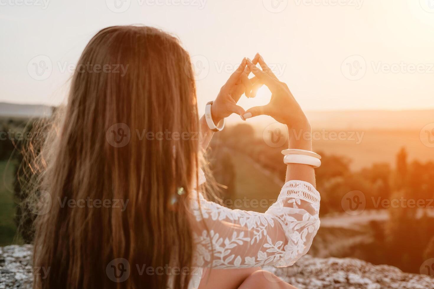 Happy woman in white boho dress making heart sign with hands on sunset in mountains. Romantic woman with long hair standing with her back on the sunset in nature in summer with open hands. photo
