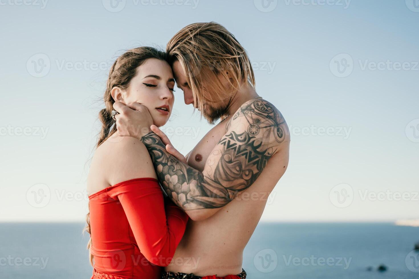 Portrait of tattooed man and sensual young brunette woman in red dress, happily dancing outdoors isolated on blurry sea background with natural bokeh in soft warm sunset backlight. Selective focus. photo