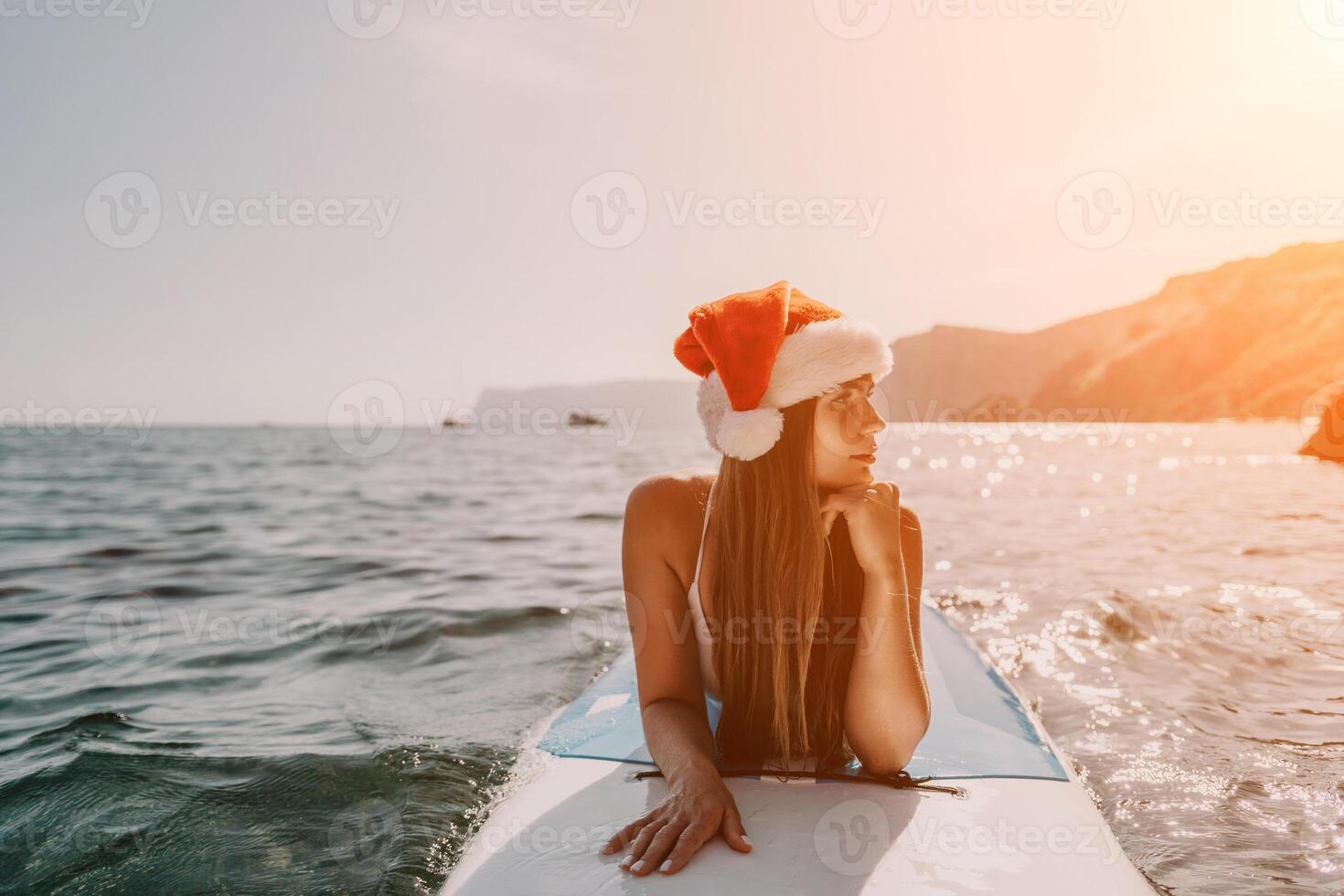 Woman sea sup. Close up portrait of happy young caucasian woman with long hair in Santa hat looking at camera and smiling. Cute woman portrait in a white bikini posing on sup board in the sea photo