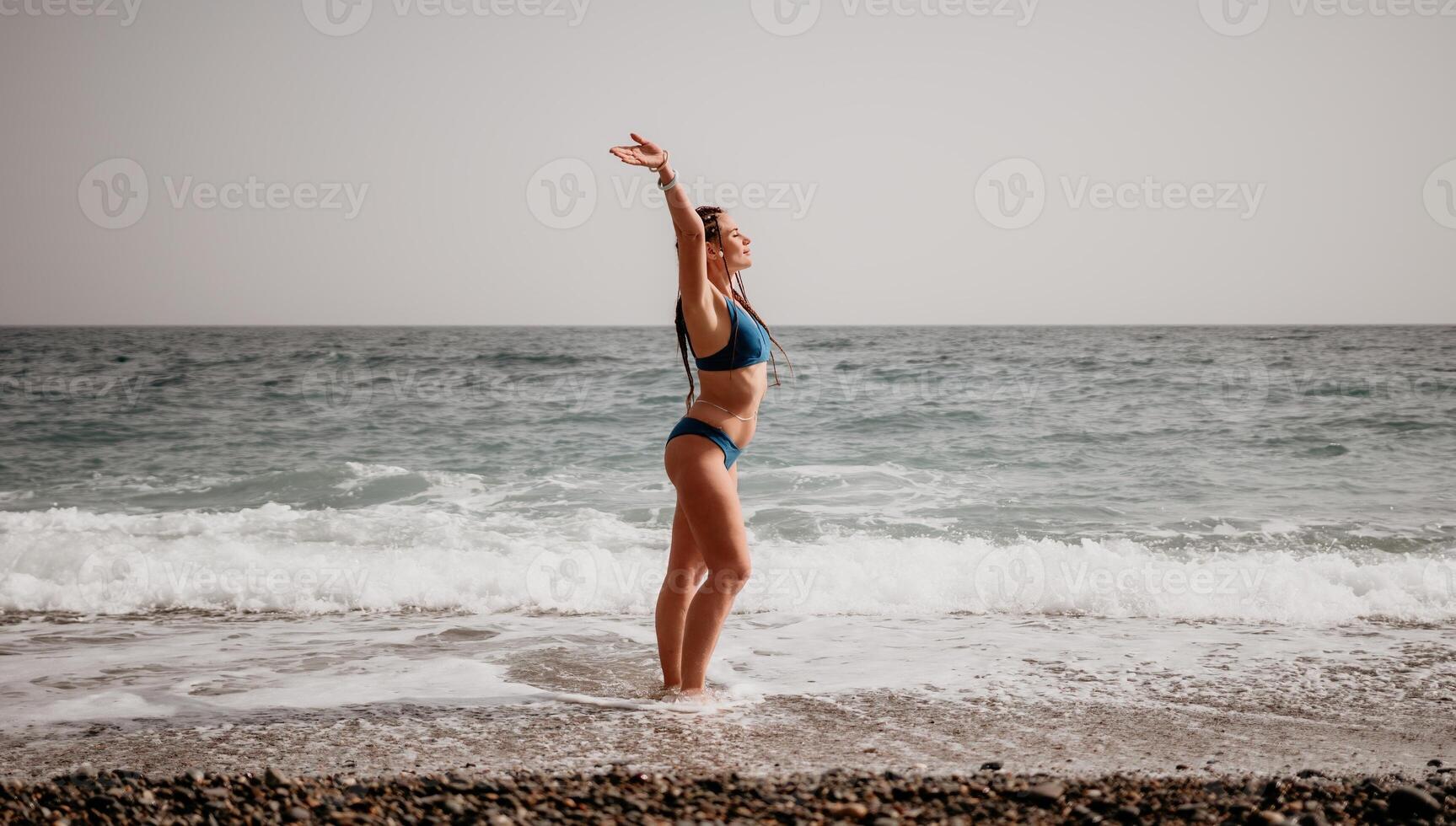 A tourist woman in blue bikini on beach enjoys the turquoise sea during her summer holiday. Rear view. Beach vacation. woman with braids dreadlocks standing with her arms raised enjoying beach ocean. photo