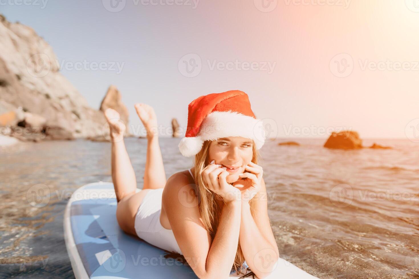 Woman sea sup. Close up portrait of happy young caucasian woman with long hair in Santa hat looking at camera and smiling. Cute woman portrait in a white bikini posing on sup board in the sea photo