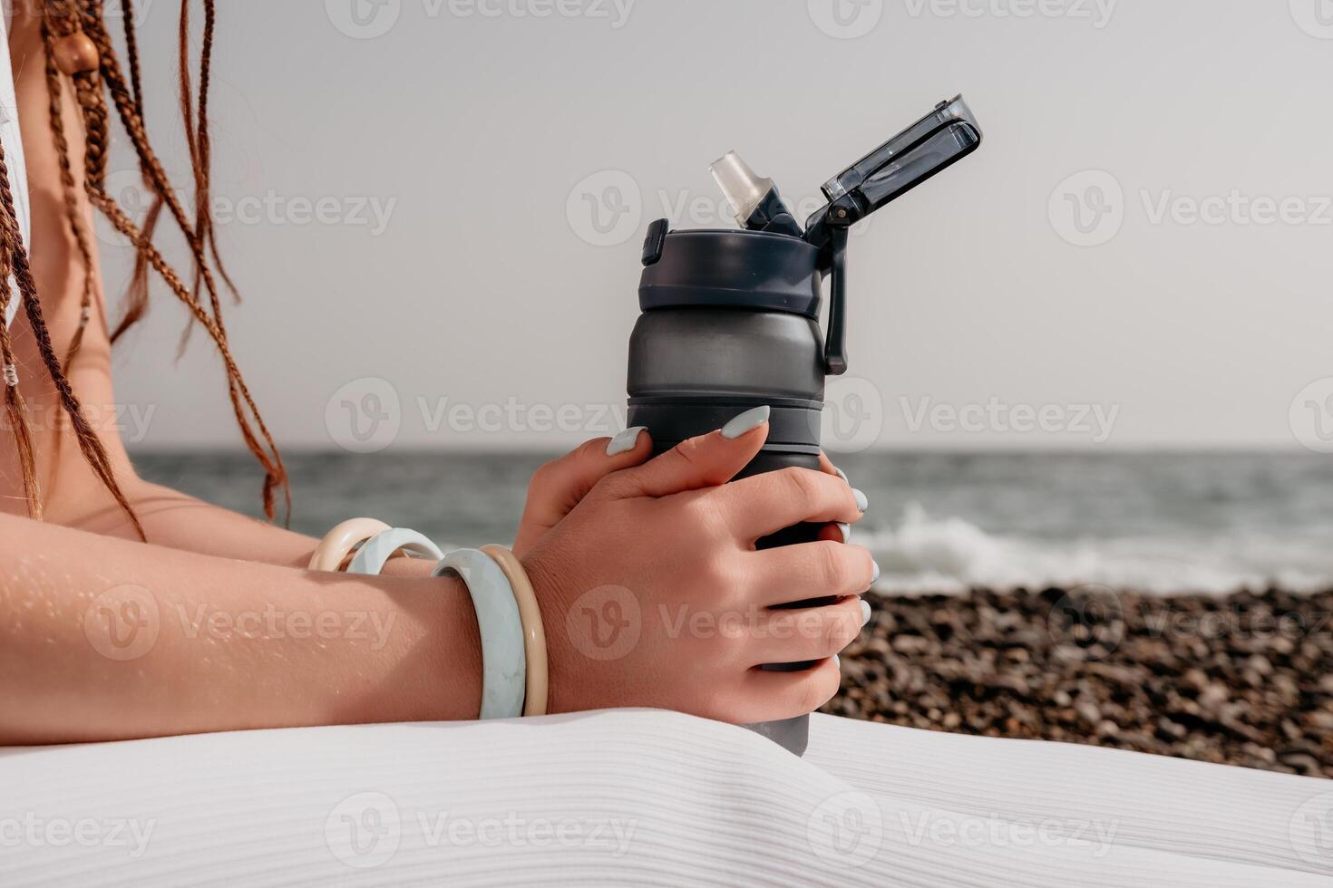 Woman sea yoga. Thermo mug in the hands of woman with braids dreadlocks in white leggings and tops doing stretching pilates on yoga mat near sea. Female fitness yoga routine concept. Healthy lifestyle photo