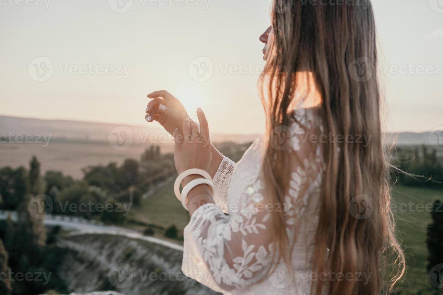 Happy woman in white boho dress on sunset in mountains. Romantic woman with long hair standing with her back on the sunset in nature in summer with open hands. Silhouette. Nature. Sunset. photo