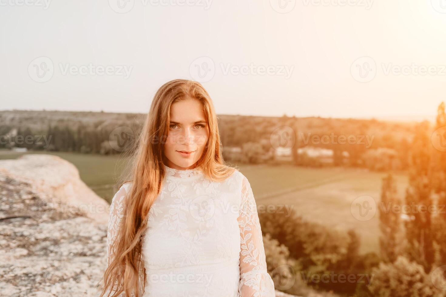 Happy woman in white boho dress on sunset in mountains. Romantic woman with long hair standing with her back on the sunset in nature in summer with open hands. Silhouette. Nature. Sunset. photo