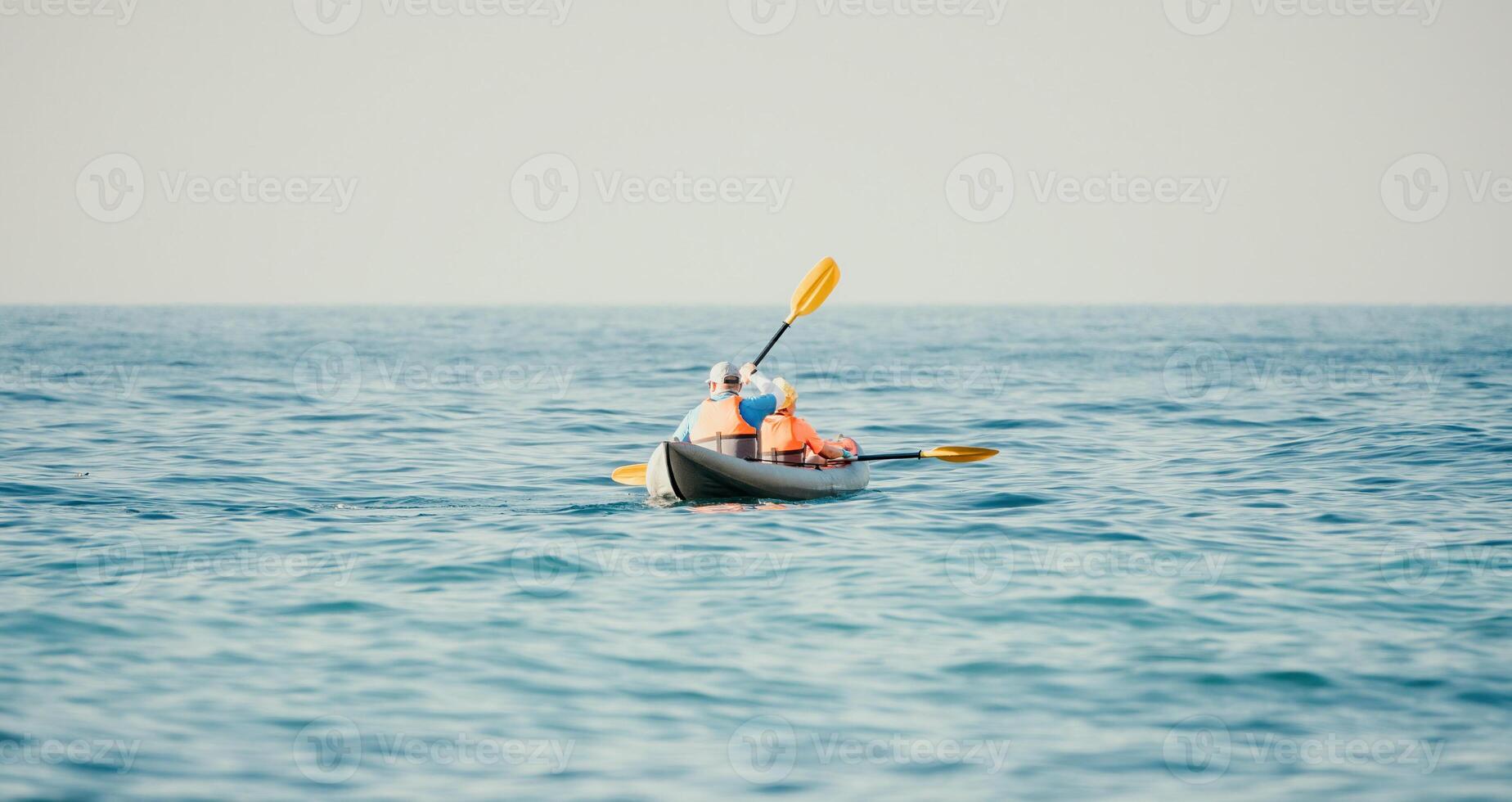 Happy couple kayaks in an inflatable kayak on the sea at sunset. Couple kanoeing in the sea near the island with mountains. People kayaking in life jackets sail. Back view photo