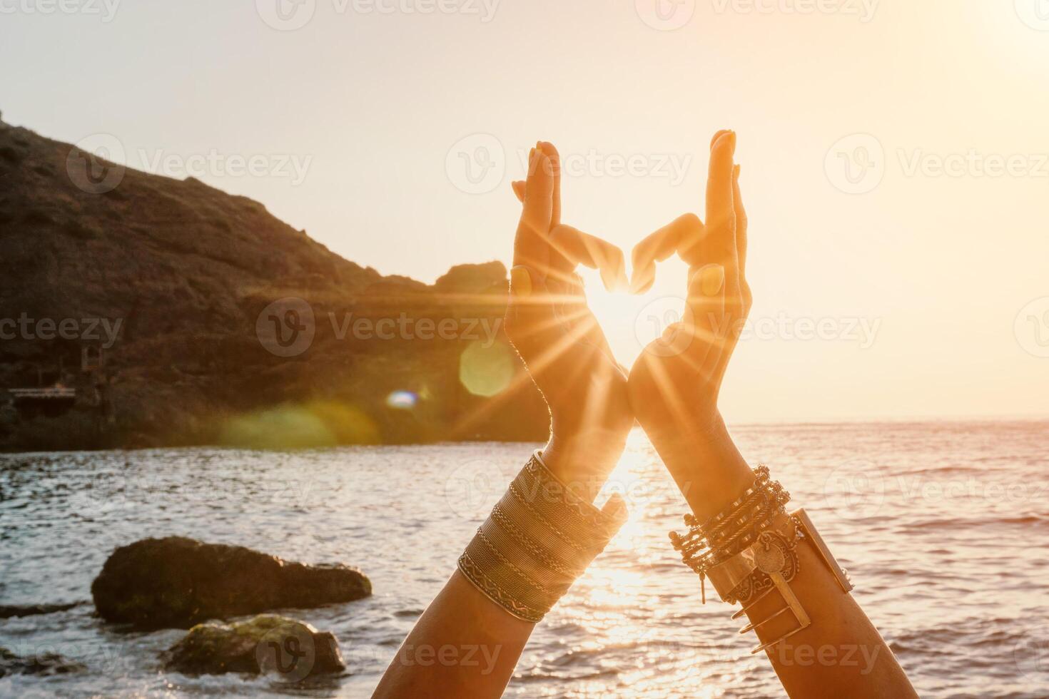 Woman sea yoga. Happy woman in a white swimsuit and boho-style bracelets making a heart shape with her hands while practicing yoga on a mat by sea at sunset. Healthy lifestyle, harmony and meditation photo