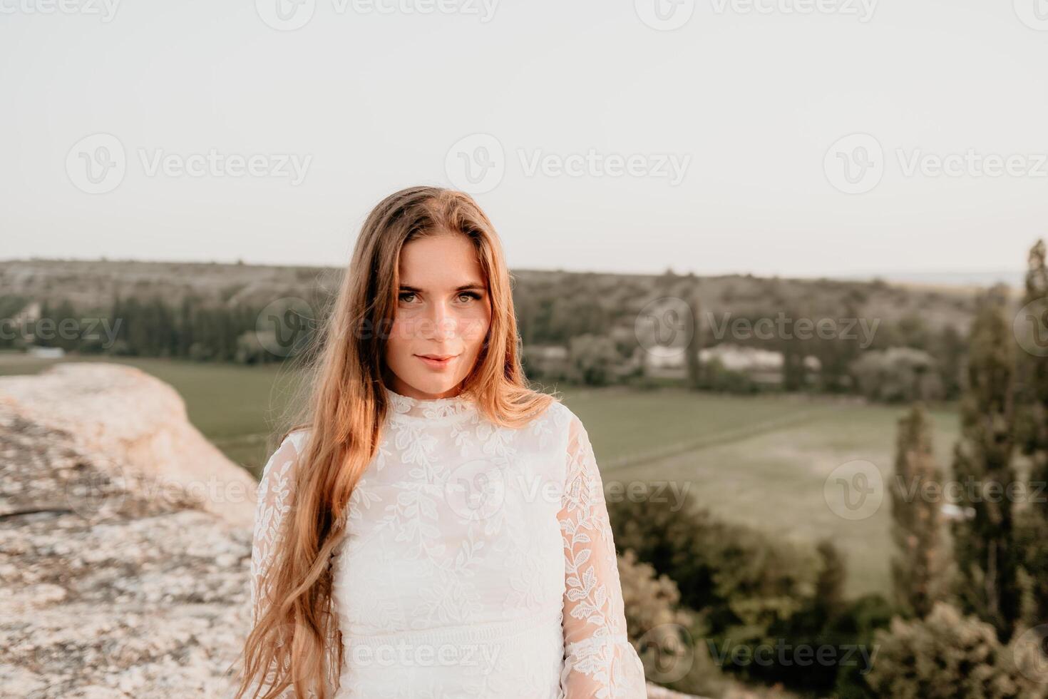 Happy woman in white boho dress on sunset in mountains. Romantic woman with long hair standing with her back on the sunset in nature in summer with open hands. Silhouette. Nature. Sunset. photo
