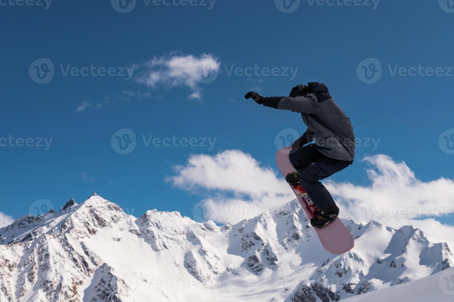 Portrait of a young man snowboarder jumping on a snowboard in sportswear, against the backdrop of high snow-capped mountains and sky photo