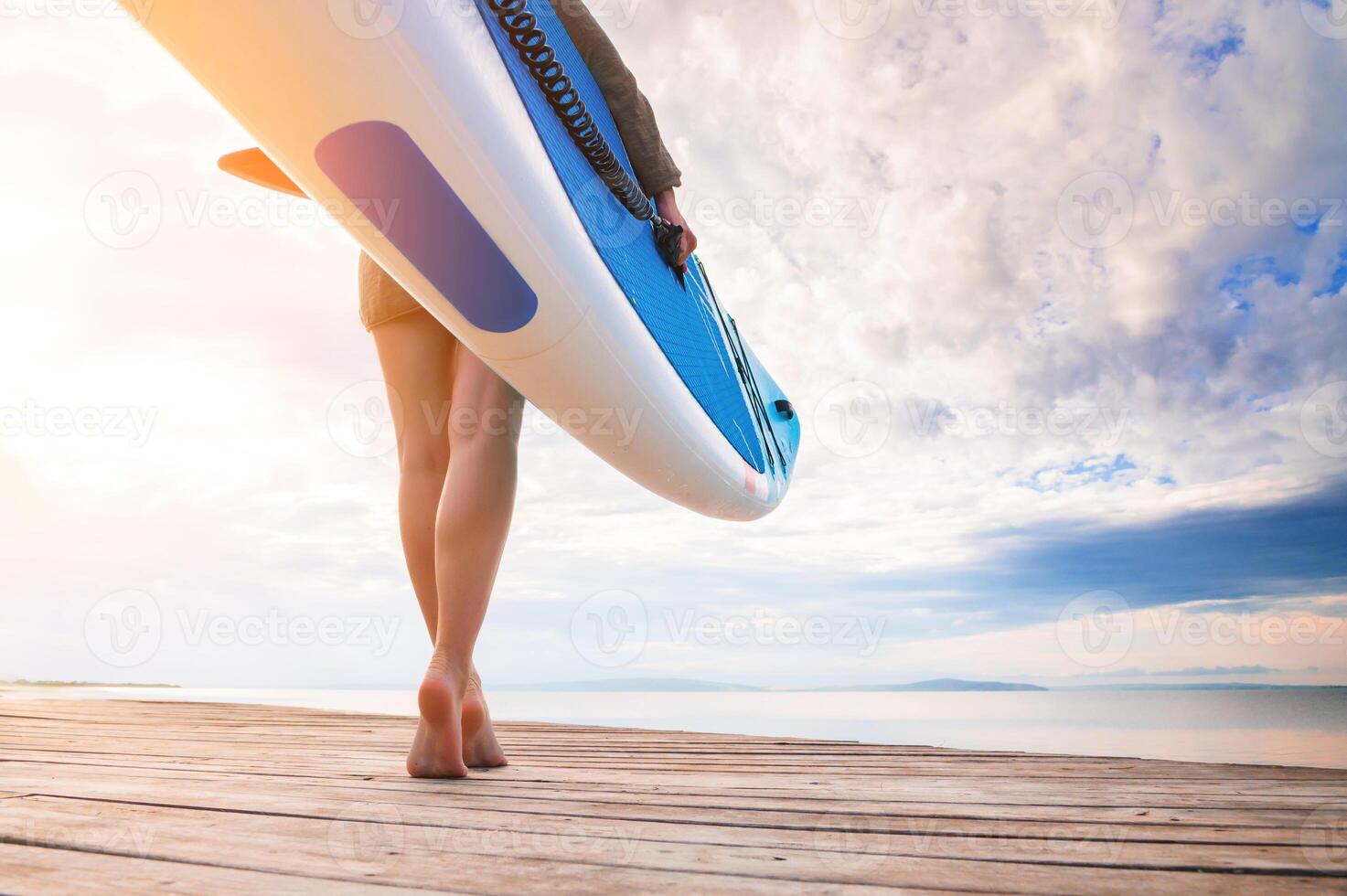 Low angle wide angle young Caucasian woman carries her paddleboard towards the water at sunset on a wooden platform. SUP and surfing concept. photo