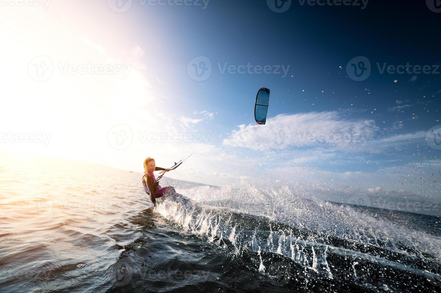 Attractive young Caucasian female kiteboarder doing a trick at sunset with splashes and a smile. Wide angle. Deep dive photo