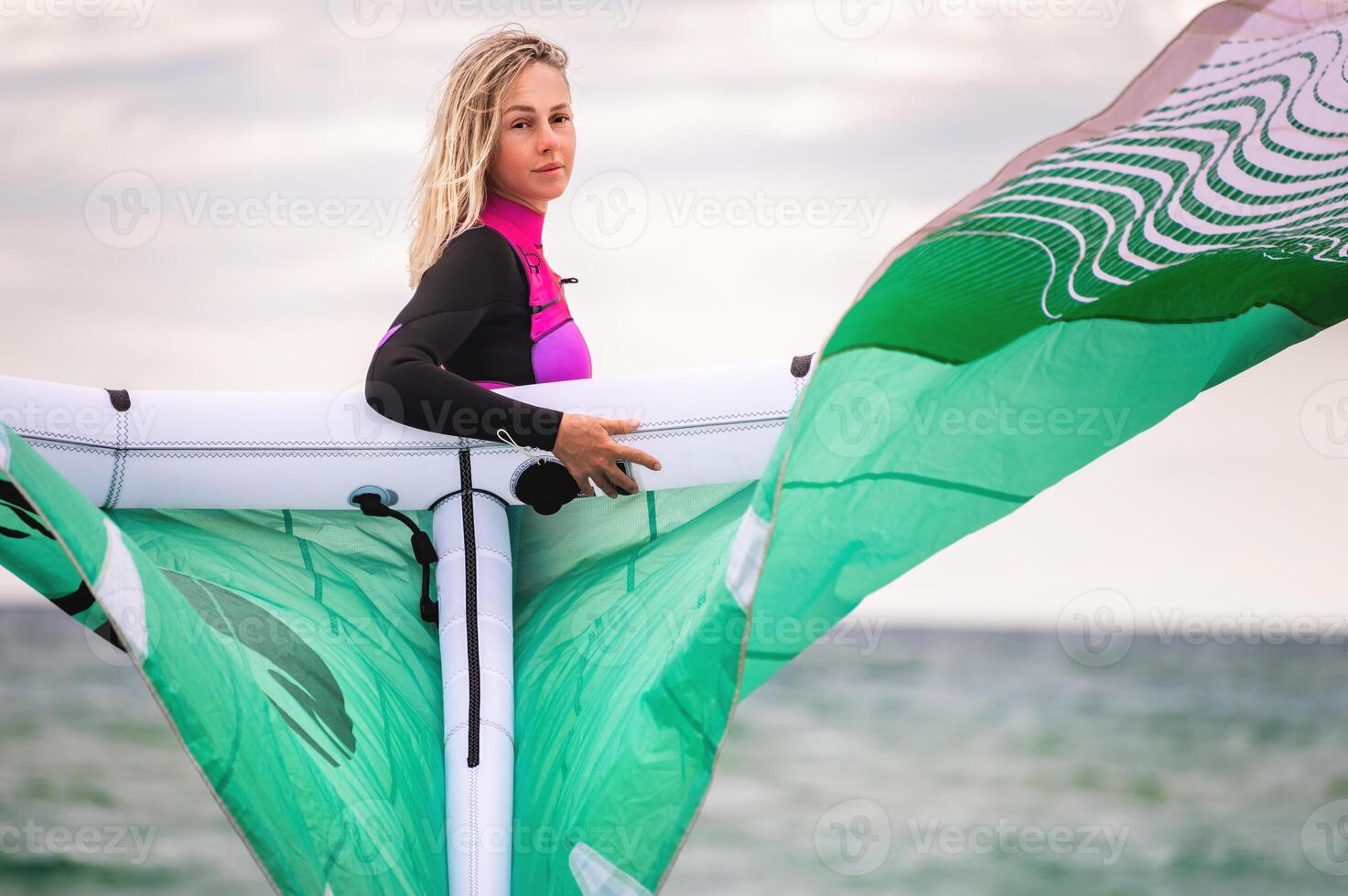 Portrait of a young female kitesurfer in a wetsuit holding her kite and looking at the camera against the background of the sea and sky photo